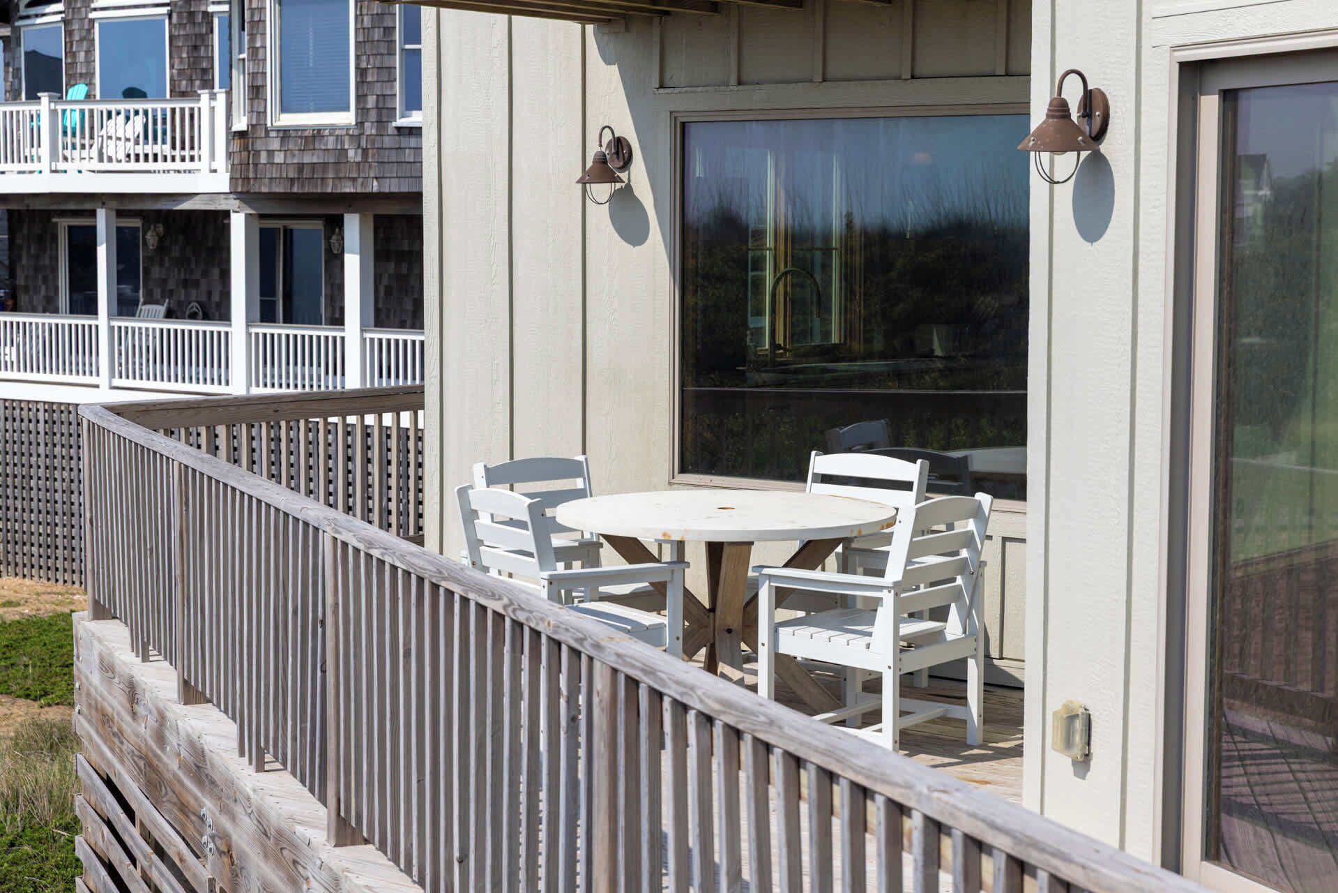 Een overdekt terras met een ronde tafel en witte stoelen, een houten hek en een gebouw met een balkon.