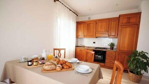Colazione servita su un tavolo in cucina, con pane, pasticcini e bevande, arredata con pensili in legno e zanzariera piastrellata.