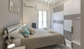 Ein Schlafzimmer mit Bett, Nachttischen, einer Chandelüster, einem Fenster und einem Kleiderschrank.