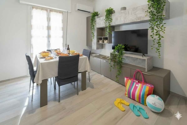 Ein Essbereich mit einem Tisch für zwei Personen, einem an der Wand befestigten Fernseher sowie einer Tasche und Flip-Flops auf dem Boden.
