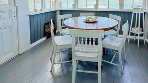 Un espace extérieur abrité, aménagé pour les repas, avec une table ronde en bois et des chaises blanches, de grandes fenêtres offrant une vue sur l’eau et un plancher en bois.