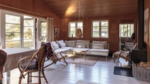 A cozy cabin interior with a wooden ceiling, large windows, a sofa, a chair, a table, and a fireplace. - Authentic summerhouse near the beach (Rørvig)