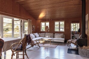 A cozy cabin interior with a wooden ceiling, large windows, a sofa, a chair, a table, and a fireplace. - Authentic summerhouse near the beach (Rørvig)