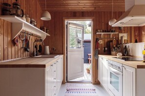 A kitchen with wooden walls, white cabinets, a large window, and a view of the outdoors. - Authentic summerhouse near the beach (Rørvig)