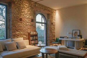 Ein gemütliches Wohnzimmer mit Steinwand, einem weißen Sofa, einem Bücherregal, einem runden Tisch und Blick durch ein großes Rundbogenfenster.