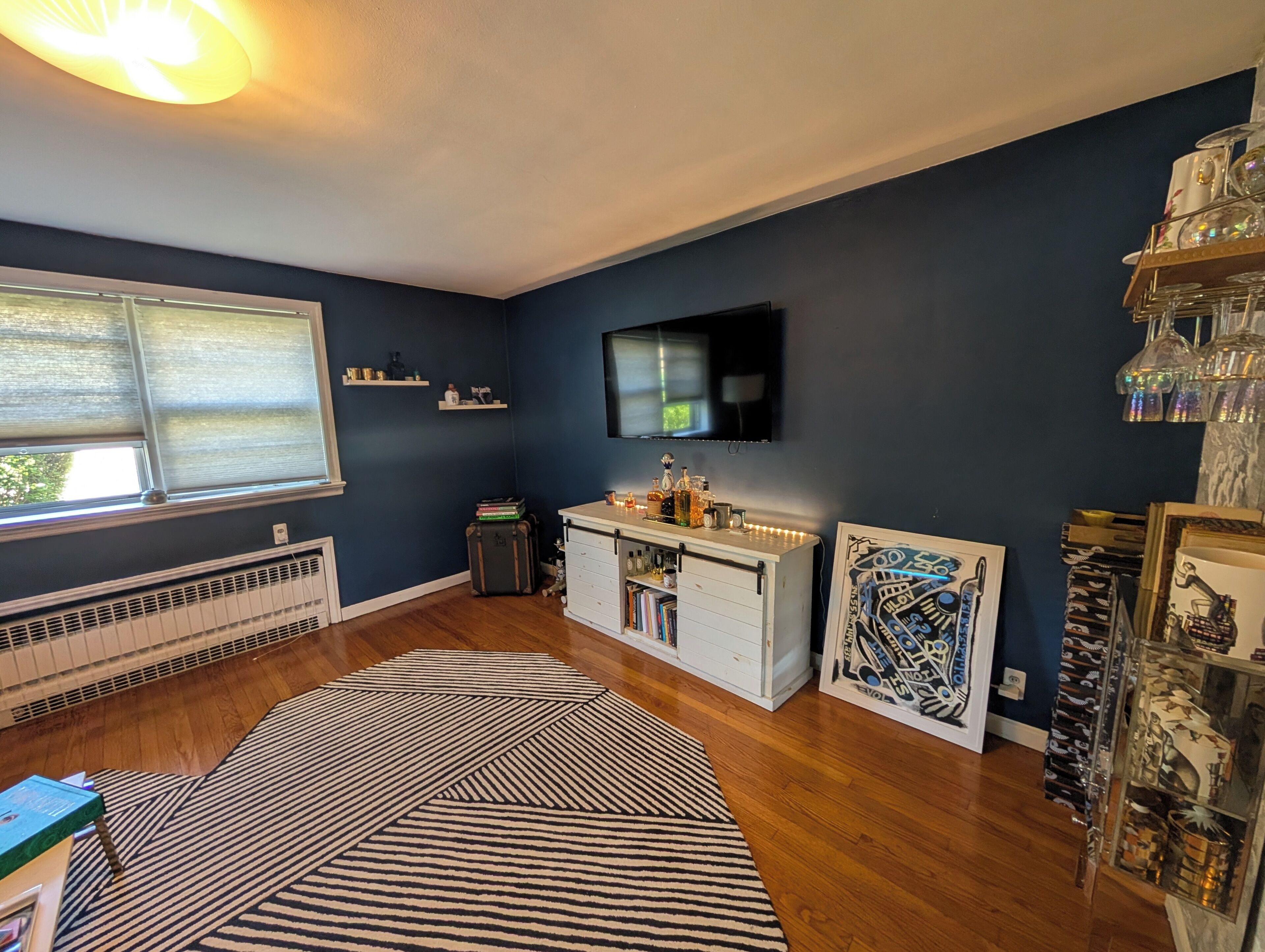 A room with a dark blue wall, a mounted TV, a white dresser with drawers, a patterned rug, and a radiator.