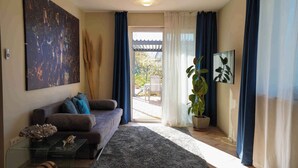 Een woonkamer met een bank, een salontafel, een groot abstract schilderij, een potplant en een uitzicht door een glazen deur op een terras.