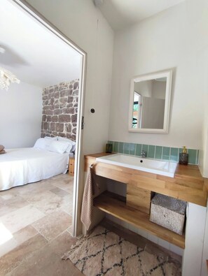 A bedroom with a stone wall, a wooden bed, and a bathroom with a wooden vanity and a mirror. - Renovated silk farm in Joyeuse (Joyeuse)