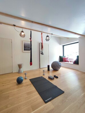 Ein geräumiges Zimmer mit Holzboden, einer schwarzen Yogamatte, Kurzhanteln, einem blauen Gymnastikball und einer Fensterbank mit Kissen.