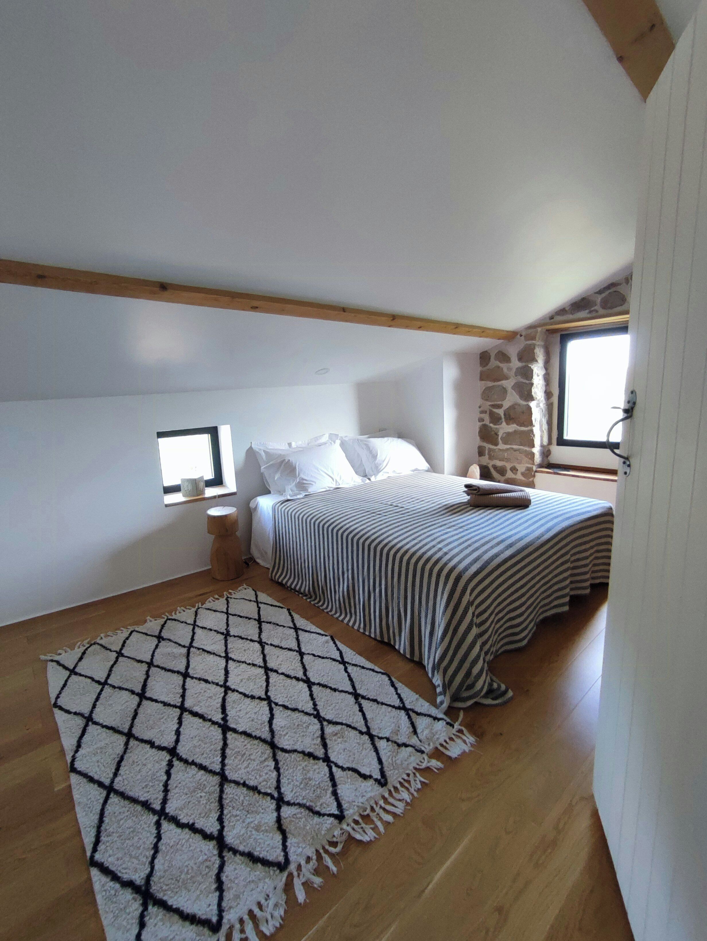 Een slaapkamer met een bed, een raam, een stenen muur en een houten balk.