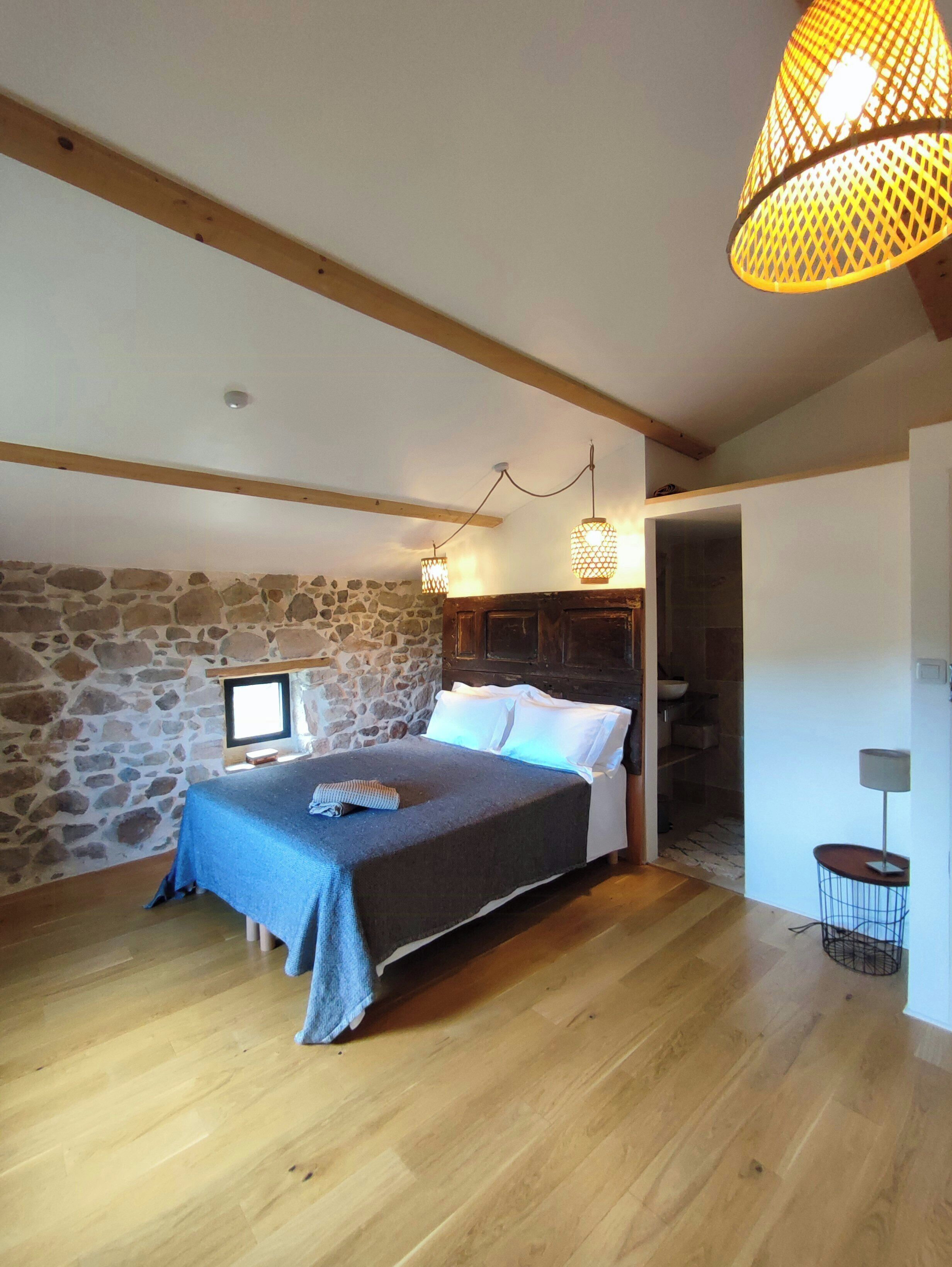 Une chambre avec un mur en pierre, des poutres apparentes, un lit recouvert de linge blanc et un luminaire suspendu.