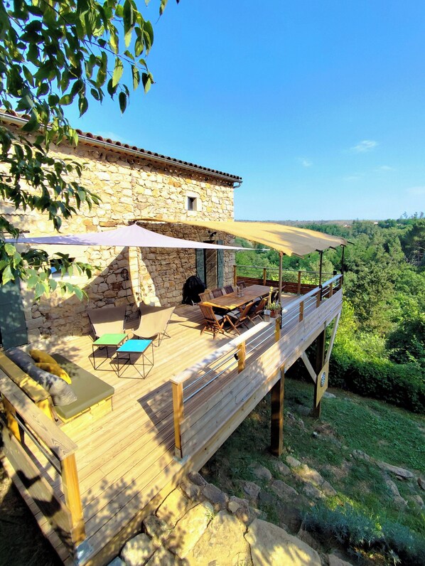 Ein Steinhaus mit einer Holzterrasse, Sitzgelegenheiten im Freien und Blick auf die umliegende Landschaft.