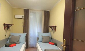 Ein Zimmer mit zwei Betten, einem Fenster mit Vorhängen, einer Klimaanlage und einer Tür.