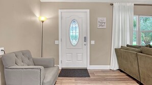 Ett vardagsrum med en beige soffa, en grå fåtölj, en vit dörr med ett fönster av färgat glas och en väggmonterad skylt.