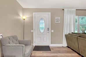 A living room with a beige sofa, a grey armchair, a white door with a stained glass window, and a wall-mounted sign. - HUGE 5bd/4ba Pool/Spa Sleeps 14+ Bunks USSSA (Melbourne)