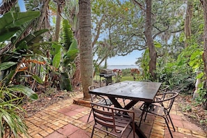 Eine Terrasse mit Tisch und Stühlen, umgeben von tropischer Vegetation und Blick auf das Wasser.