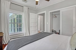 Ein Schlafzimmer mit Bett, einem Fenster mit Vorhängen, einer Tür, einem Heizkörper und einem Kleiderschrank mit Kleiderstange.