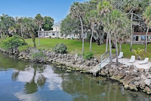 Ein Wohnhaus direkt am Wasser mit einem Steg, Liegestühlen und einem großen Haus im Hintergrund.