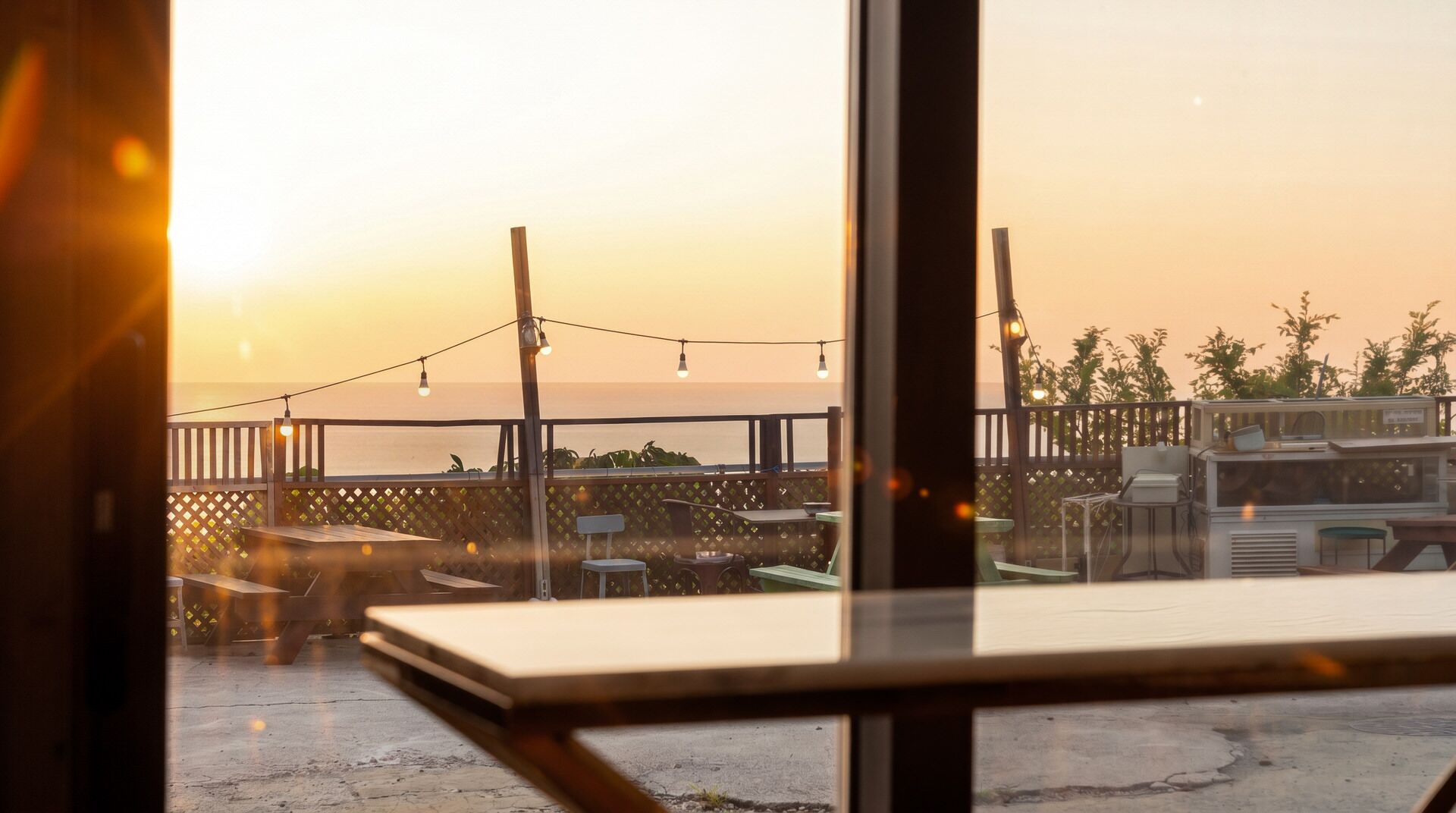A view from inside a building looking out to a patio area with tables and chairs, a grill, and a fence overlooking a body of water.