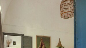Ein Innenraum mit einer Tür zu einem anderen Raum, einer Bank mit Büchern und einem Ventilator, einem Wandbild eines Segelbootes und einem hängenden Vogelkäfig.