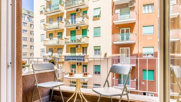 En balkon med to stole og et lille bord, med udsigt over en bygning med flere etager, altaner og vinduer.