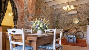 Un comedor rústico con una mesa de madera, sillas blancas, un jarrón de flores, una bicicleta y un reloj en la pared.