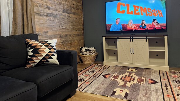 A living room with a black sofa, a patterned throw pillow, a flat-screen TV displaying 'Clemson,' a wooden wall, and a patterned rug. - CountryComfortRetreat!!! 2-bedroom mobile home with AC in Birch Tree (Birch Tree)
