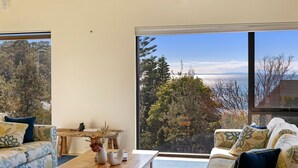 Een kamer met een groot raam dat uitzicht biedt op bomen en een wateroppervlak. Er staat een houten tafel met een vaas en een camera.