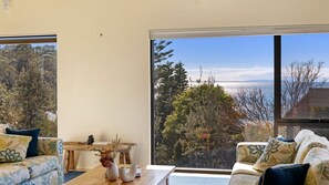 Ein Zimmer mit großem Fenster und Blick auf Bäume und ein Gewässer. Es steht ein Holztisch mit einer Vase und einer Kamera darauf.