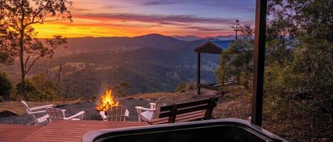 A hot tub with a fire, two chairs, and a bench on a wooden deck with a view of mountains and a sunset.