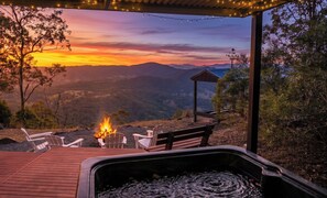 A hot tub with a fire, two chairs, and a bench on a wooden deck with a view of mountains and a sunset.