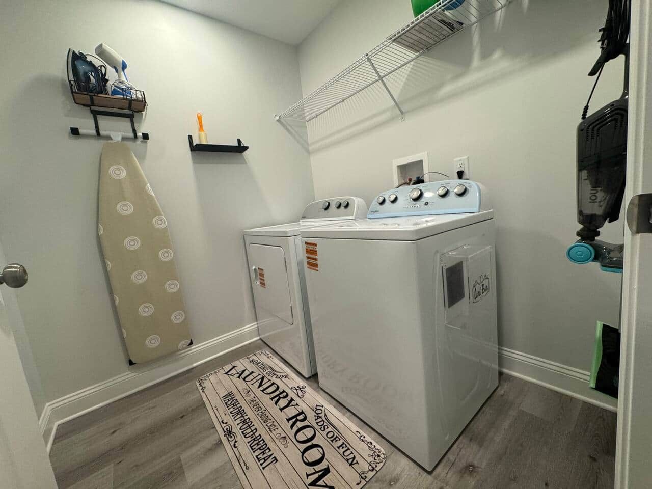 A laundry room with a washing machine, dryer, ironing board, and a shelf with laundry supplies.
