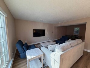 Uma sala de estar com sofá, poltronas, mesa de centro, televisão de tela plana na parede e um espaço de jantar com mesa e cadeiras.