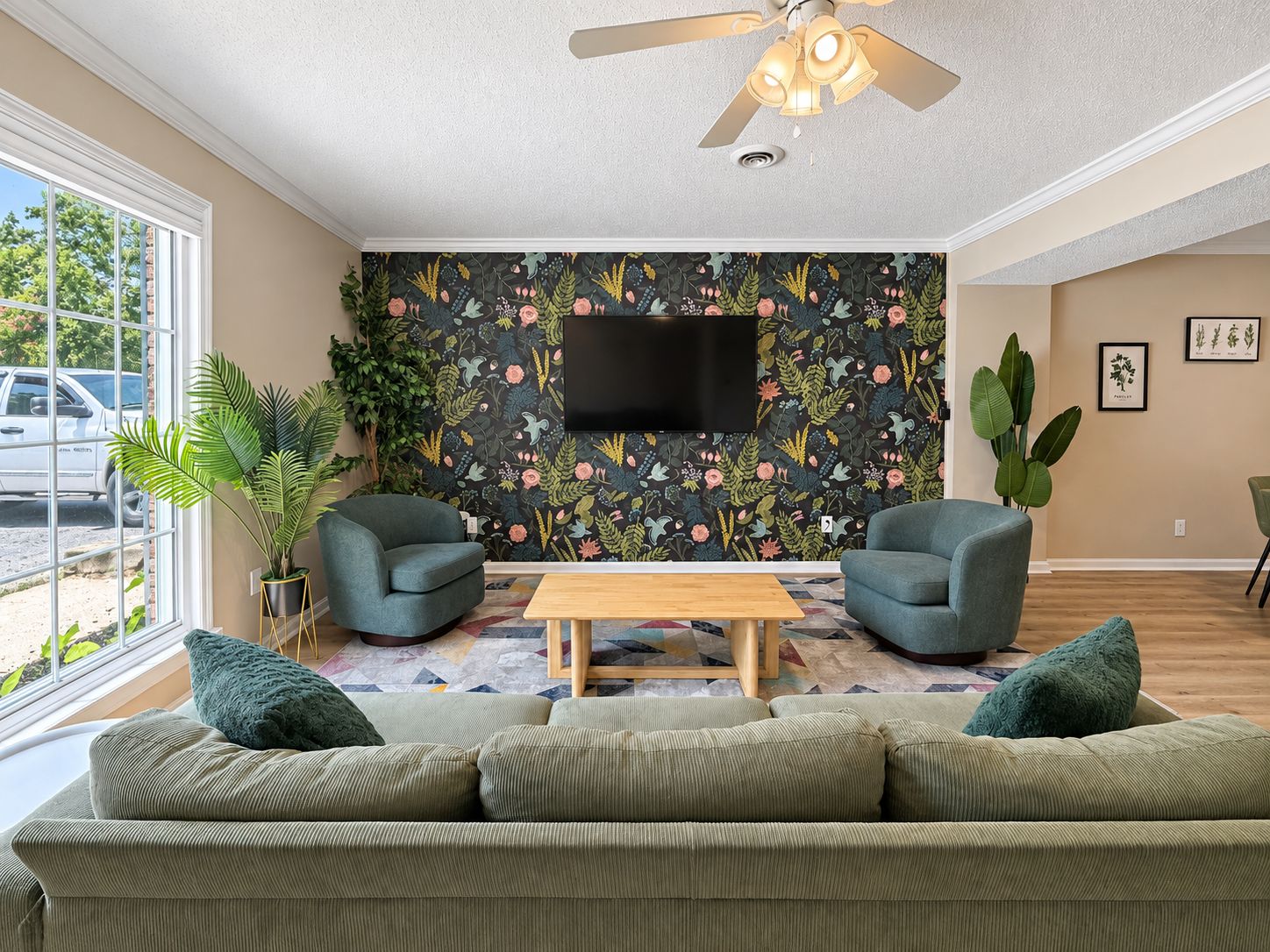 Un salon comprenant un canapé, deux fauteuils, une table basse, un ventilateur de plafond, un téléviseur et un papier peint à motifs floraux.