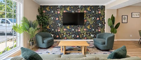 Un saló amb un sofà, dues butaques, una taula de centre, un ventilador de sostre, un televisor i un fons de pantalla amb motius florals.