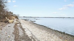 Ein Strand mit einem hölzernen Steg, der sich ins ruhige Wasser erstreckt, umgeben von spärlicher Vegetation und wolkenlosem Himmel.