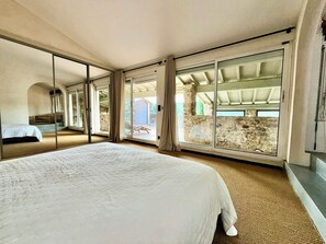 Ein Schlafzimmer mit einem großen Bett, Schiebeglasfenstern zu einem Außenbereich und einer dahinter sichtbaren Steinwand.