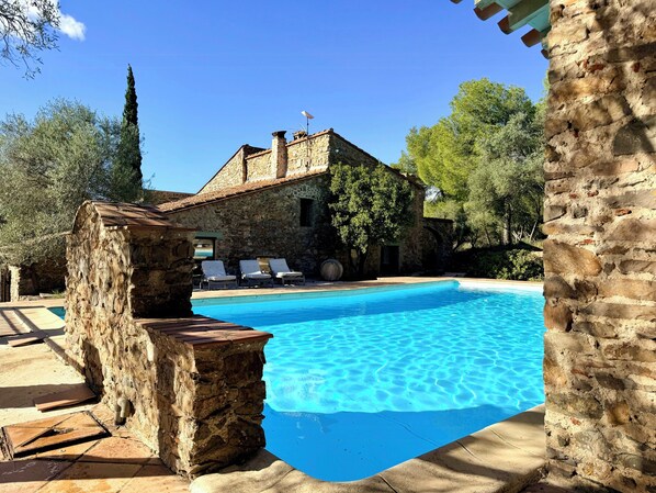 Ein Steinhaus mit Swimmingpool, Liegestühlen und einem Baum.