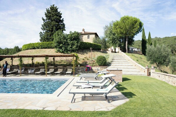 Area piscina con lettini, un vialetto in pietra e un edificio sullo sfondo.