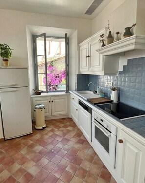 A kitchen with white cabinets, a window with a view of pink flowers, a trash bin, and a stove. - Village house with valley view in Tourrettes (Tourrettes)
