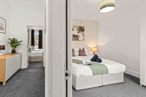 Habitación de hotel con cama, mesita de noche, lámpara y vista a otra habitación.