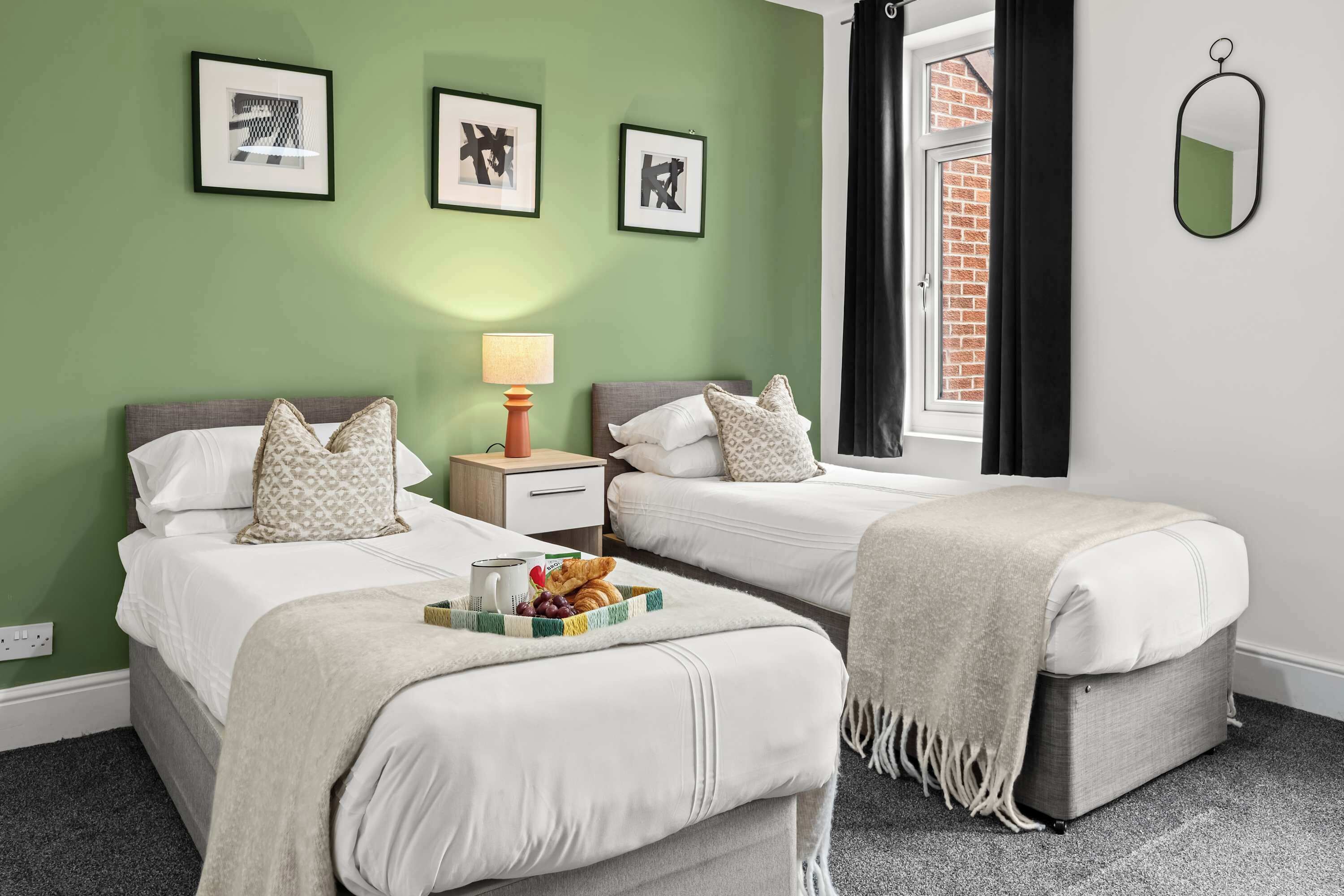 A hotel room with two beds, a small table with a lamp, and a window with a view of a brick building.