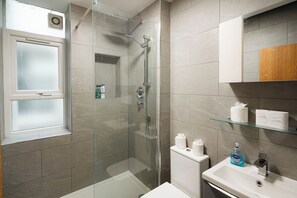 A modern bathroom with a glass shower enclosure, a toilet, a sink with a mirror, and a window. - Modern 3BR Flat in Chelsea (London)
