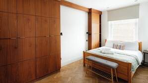 A bedroom with a wooden wardrobe, a bed with a bench, and a window with blinds. - Modern 3BR Flat in Chelsea (London)