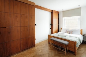 A bedroom with a wooden wardrobe, a bed with a bench, and a window with blinds. - Modern 3BR Flat in Chelsea (London)