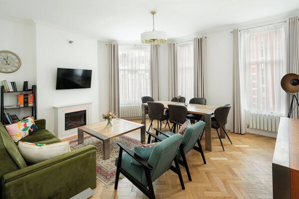 A modern living room with a fireplace, a television, a dining area with chairs, and large windows with curtains. - Modern 3BR Flat in Chelsea (London)