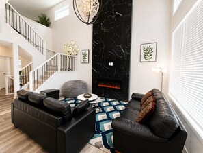 Een moderne woonkamer met een open haard, zwarte leren banken, een trap en een marmeren wand.