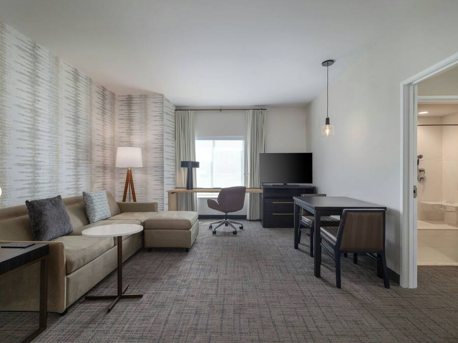 A hotel room with a sofa, a small table, a desk, a chair, a TV, and a bathroom visible through an open door.