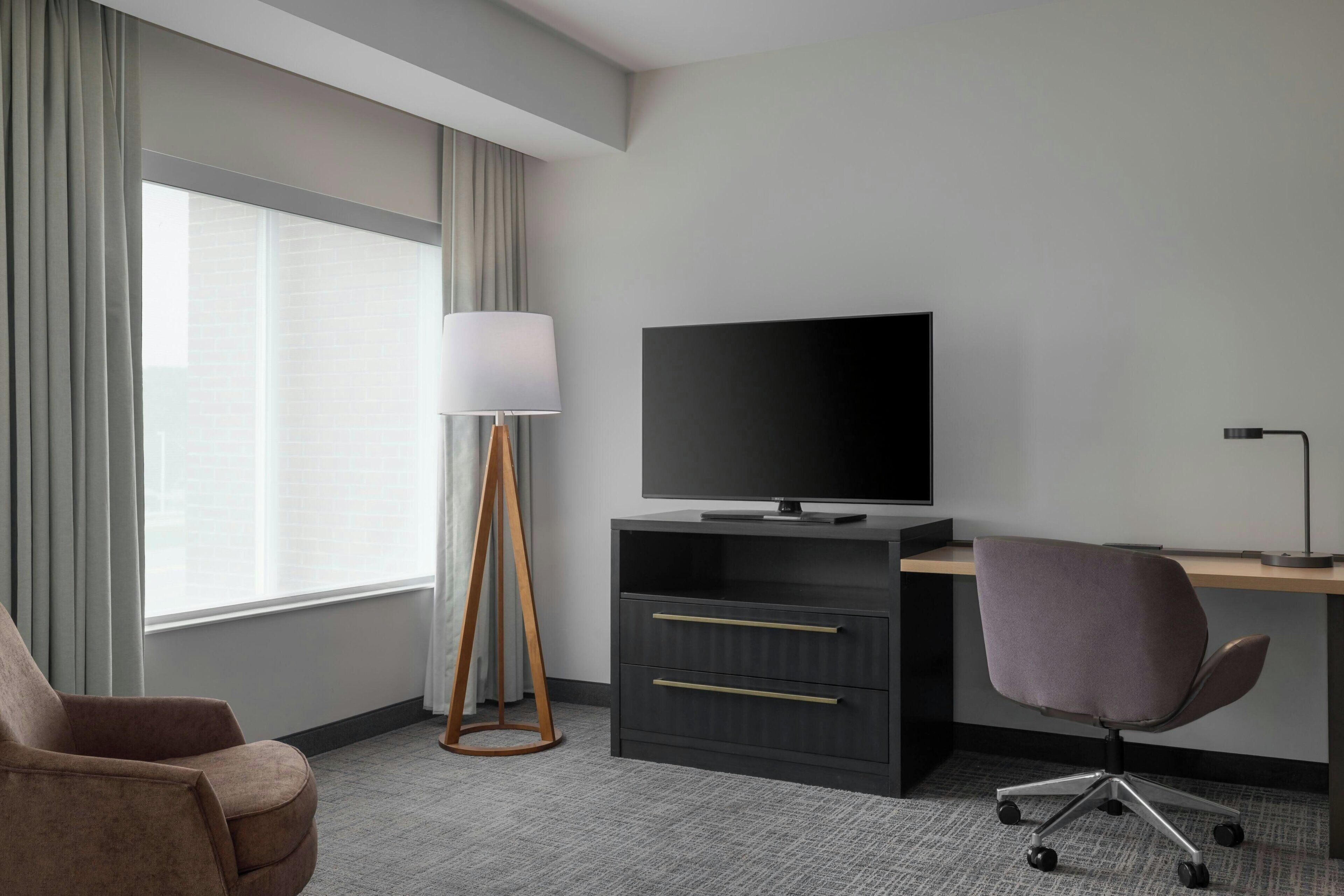 A hotel room with a desk, chair, television, lamp, and a large window with curtains.