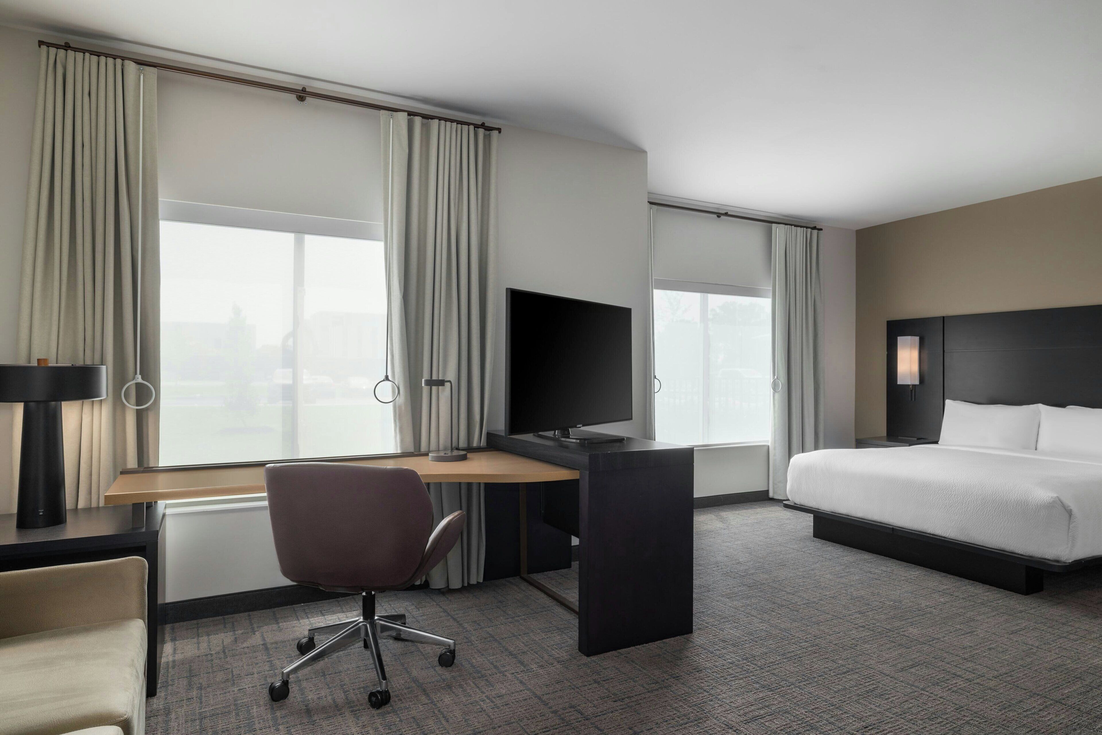 Habitación de hotel con cama, escritorio, silla, televisor y ventanas grandes con cortinas.